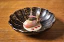 A decorative dark bowl with a piece of raw fish topped with caviar, a lemon slice, and a crisp garnish, served on a creamy white sauce with pink sprigs and pomegranate seeds, on a wooden table.