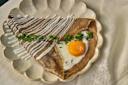 A folded buckwheat crepe topped with a sunny-side-up egg, drizzled with white sauce, and garnished with chopped fresh herbs, served on a scalloped ceramic plate over a light tablecloth.