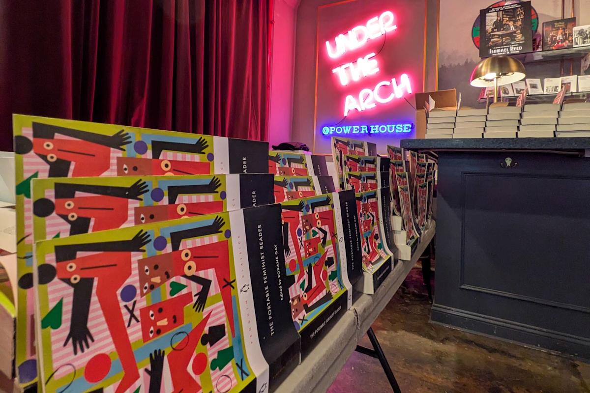 A table displays colorful, abstract art books in front of a neon sign reading "UNDER THE ARCH @ POWERHOUSE" inside a bookstore with stacked books and dark curtains in the background.