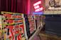 A table displays colorful, abstract art books in front of a neon sign reading "UNDER THE ARCH @ POWERHOUSE" inside a bookstore with stacked books and dark curtains in the background.