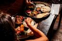 Two people sitting at a dark wooden table, enjoying Indian cuisine served on metal plates with rice, curries, naan, papadum, and various colorful side dishes. One person is reaching for food with their hand.