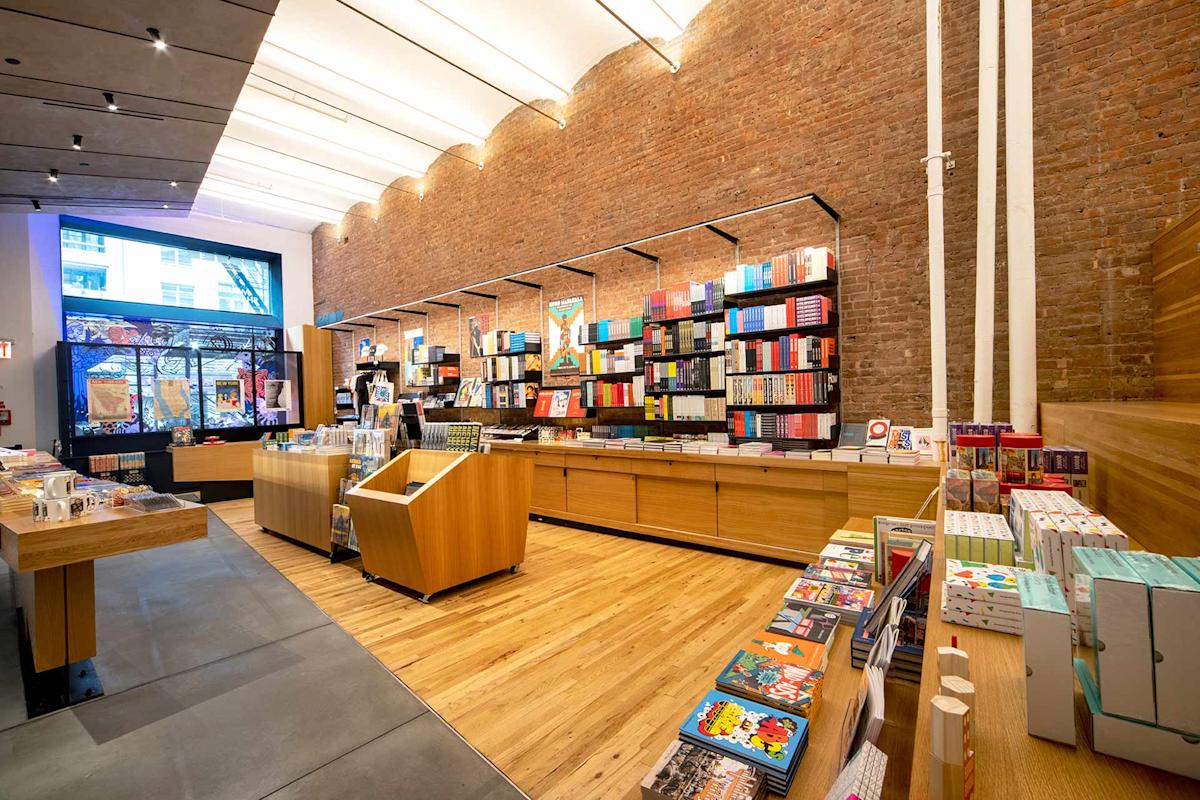 A modern bookstore with wooden shelves and tables displaying colorful books and gifts, large windows letting in natural light, and exposed brick walls creating a cozy atmosphere.
