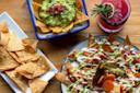 Colorful Mexican meal featuring nachos topped with sauces and pickled vegetables, a bowl of guacamole with tortilla chips, extra chips on a plate, and a pink drink garnished with a sprig of rosemary on a wooden table.