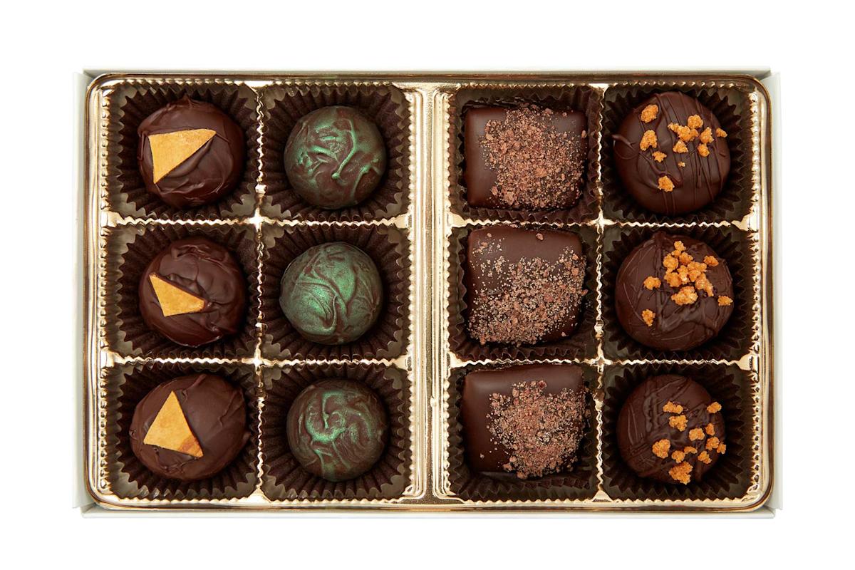 A box of twelve assorted gourmet chocolates, each in a brown paper cup, featuring various toppings and designs, including gold triangles, green swirls, cocoa dusting, and caramelized bits.