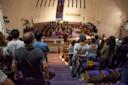People are standing in pews inside a church, facing the altar where a choir is singing. The church has purple seating and a prominent stained glass window. Some attendees are hugging while others watch the performance.