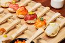 A wooden tray holds wooden spoons, each topped with a bite-sized portion of sushi, including various types of fish and garnishes, arranged neatly in rows.