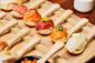A wooden tray holds wooden spoons, each topped with a bite-sized portion of sushi, including various types of fish and garnishes, arranged neatly in rows.