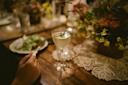 A hand holds a fork near a plate of salad on a wooden table, which is decorated with a lace doily, flowers, and a cocktail garnished with herbs. The scene has warm, ambient lighting.