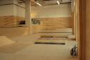 Indoor skate park with wooden ramps, platforms, and a fence. A skateboard rests against the wall. The space has high ceilings and bright lighting, creating an open environment for skateboarding activities.