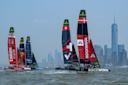 Sailboats with colorful sails race on the water with the New York City skyline and One World Trade Center visible in the background under a partly cloudy sky.