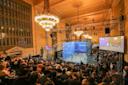 A squash game in a grand indoor arena with spectators seated around a glass court. Chandeliers and ornate architectural details adorn the venue. An audience watches intently, with a large screen displaying the game on the right.