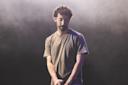 A man with curly hair and a beard stands on stage in a gray t-shirt, holding a microphone and looking down, with a foggy, dark background behind him.