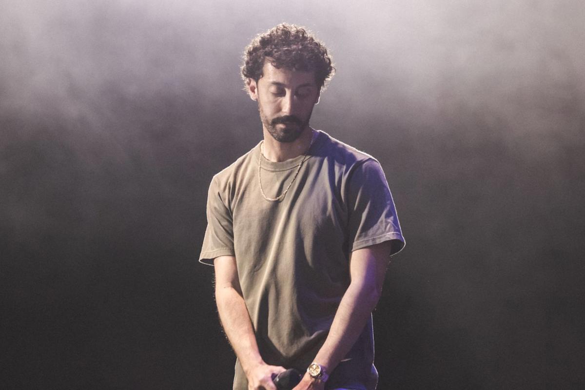 A man with curly hair and a beard stands on stage in a gray t-shirt, holding a microphone and looking down, with a foggy, dark background behind him.