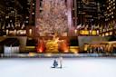 A person kneels, proposing to another person on an ice rink at Rockefeller Center. A large, brightly lit Christmas tree and a golden statue are in the background. The scene is framed by surrounding buildings and flags.