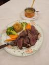 A plate with sliced steak, broccoli, carrot sticks, and a mound of seasoned rice, garnished with herbs. A small pot of creamy sauce sits on a separate plate in the background.