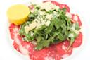 Thinly sliced raw beef topped with arugula and shaved Parmesan on a white plate. A lemon half sits to the side, enhancing the presentation of this classic carpaccio dish.