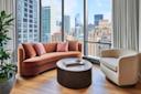 A modern living room with a curved brown sofa, a round black coffee table, and a beige armchair. Large windows offer a view of a city skyline with various buildings. A potted plant sits in the corner on wooden flooring.