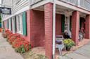 A brick building with green shutters, vibrant red flowers lining the sidewalk, and gray Adirondack chairs with a pot of yellow flowers on a porch under a wooden overhang.