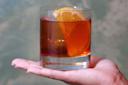 A hand holds a glass of dark brown cocktail garnished with a large orange slice, against a blurred, muted background.