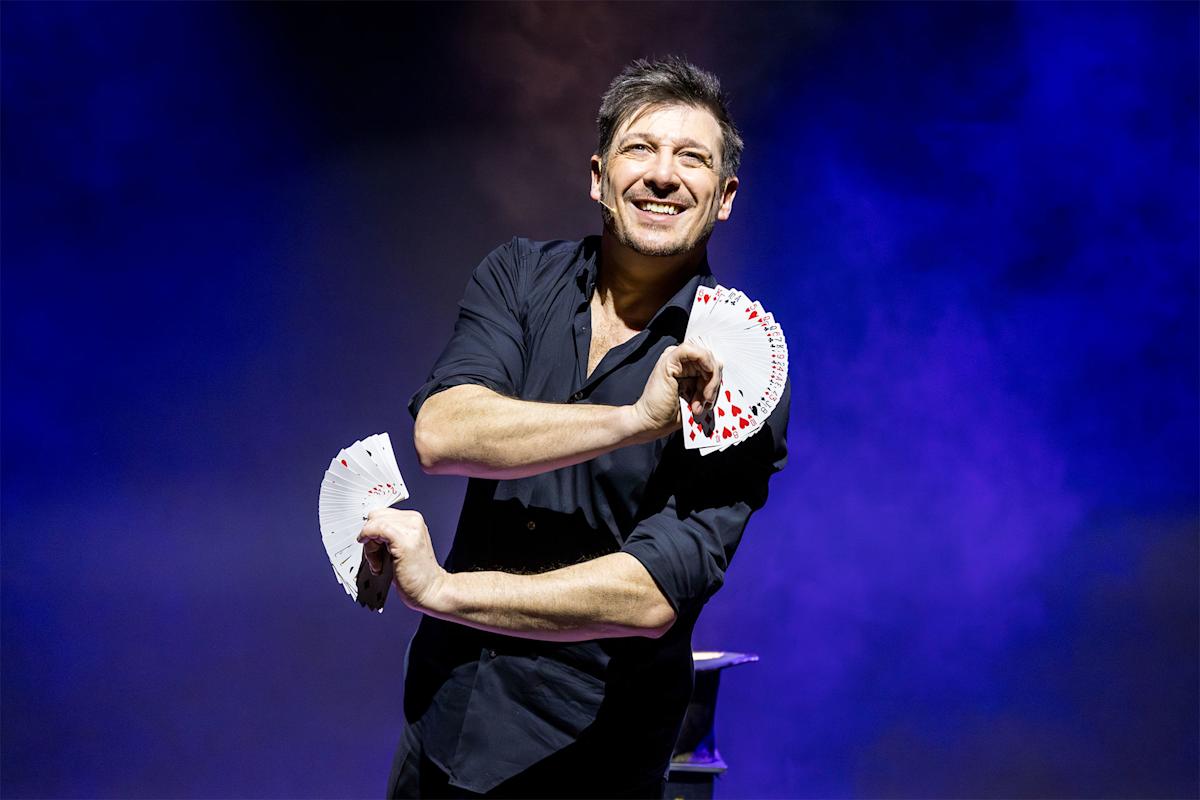 A smiling magician in a dark shirt holds two fanned decks of cards against a dramatic purple-lit background, performing a card trick.