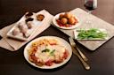 A meal setting featuring a plate of breaded chicken topped with cheese and tomato sauce, served with spaghetti. Surrounding dishes include stuffed balls on tomato sauce, green beans, dessert pastries, and a glass of red wine on a stone platter.