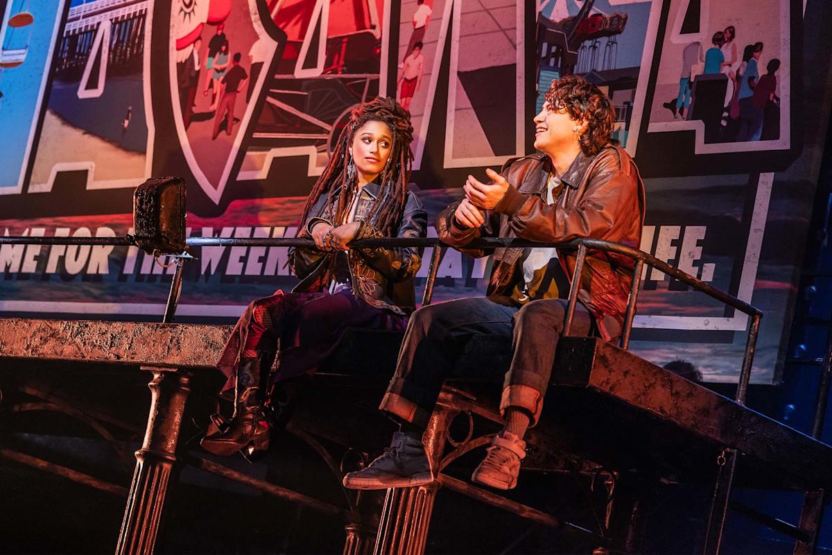 Two actors sit on a raised platform on stage, talking and gesturing. Behind them is a large, colorful postcard-style sign with images and text. The lighting is warm, creating a dramatic theater scene.