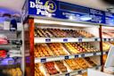 A display case at The Donut Pub shows rows of assorted donuts, crullers, and pastries. Shelves are filled with colorful donuts, and merchandise like hats and shirts are visible on the left. Blue menu boards hang above.