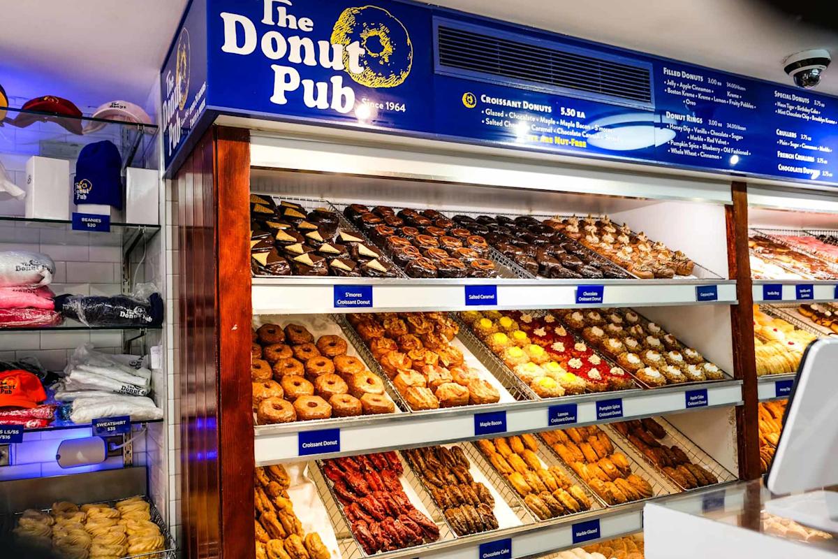 A display case at The Donut Pub shows rows of assorted donuts, crullers, and pastries. Shelves are filled with colorful donuts, and merchandise like hats and shirts are visible on the left. Blue menu boards hang above.