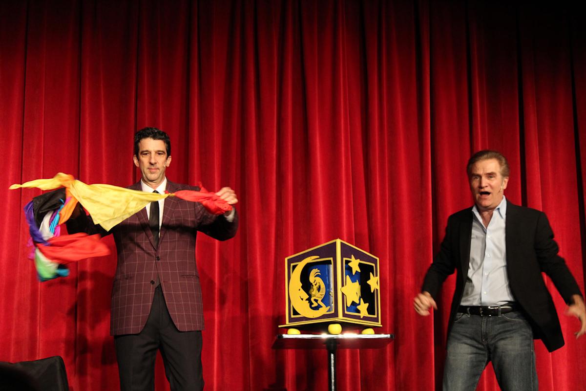 Two men perform a magic trick on stage in front of red curtains; one man holds colorful scarves, while the other gestures beside a decorative box on a small round table.