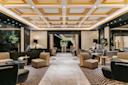 Luxurious hotel lobby featuring modern armchairs, sofas, and low tables on patterned carpets, with marble flooring, decorative ceiling panels, soft lighting, and plants adding a touch of greenery.