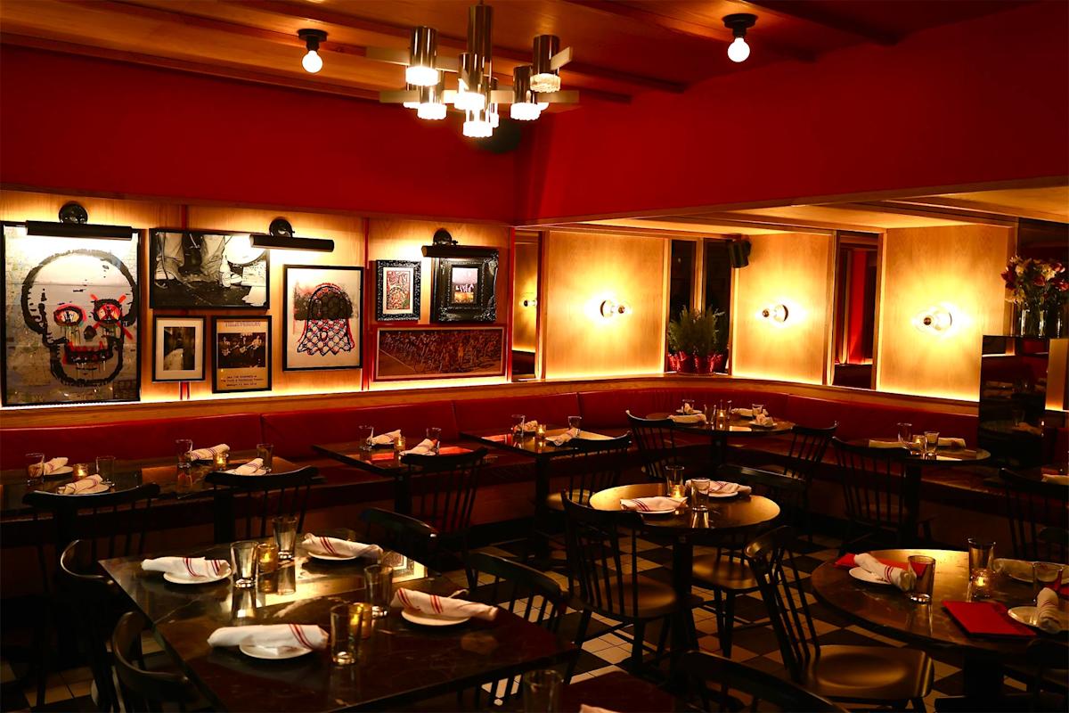 A warmly lit restaurant with red walls, modern lighting, black chairs, and tables set with glasses and napkins. Artworks, including a skull and basketball hoop, decorate the walls. The atmosphere is cozy and inviting.