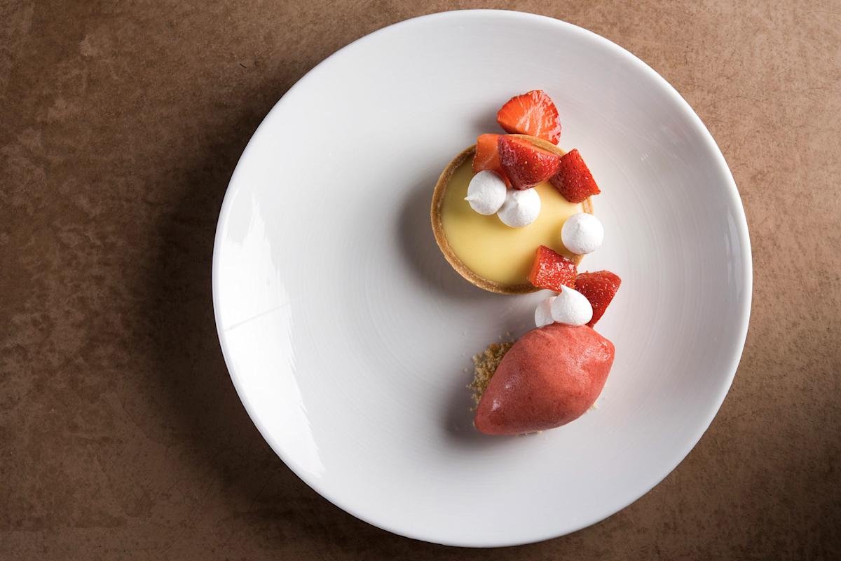 A white plate with a lemon tart topped with strawberries and small meringues, next to a scoop of strawberry sorbet, set on a brown textured surface.