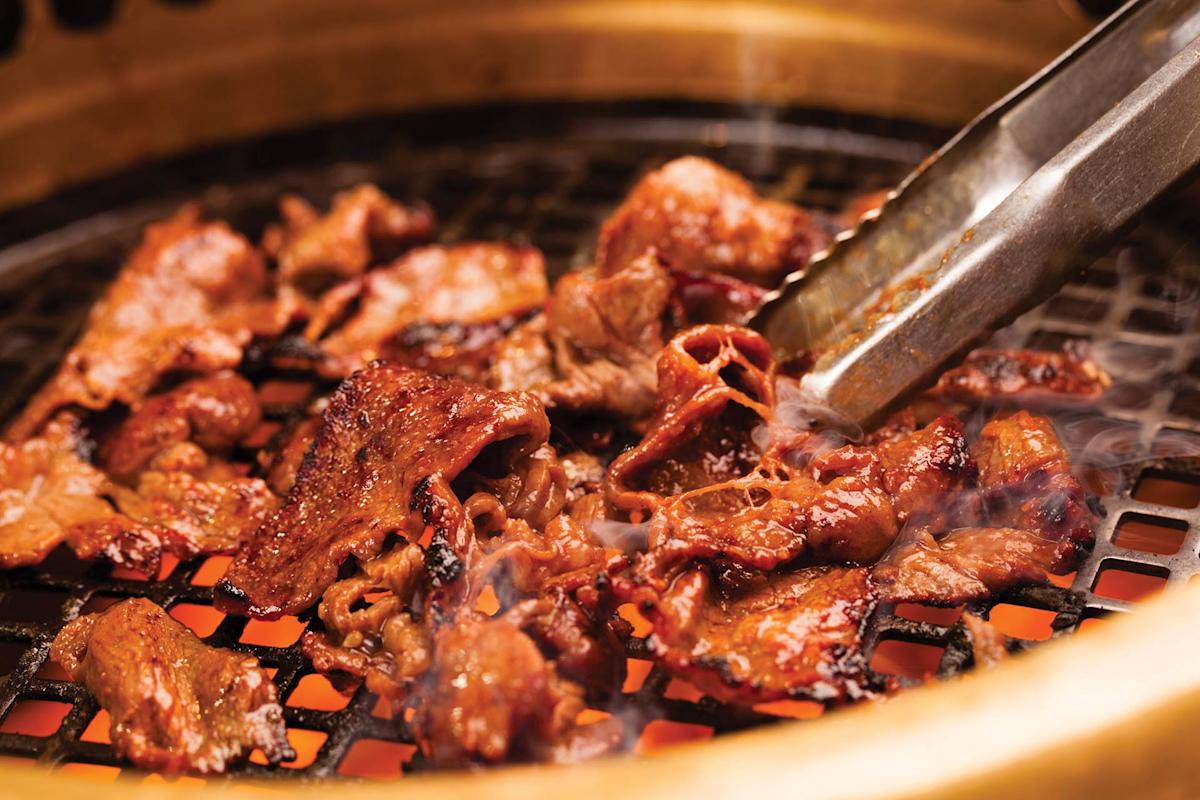 Slices of marinated meat are being grilled over a charcoal grate, with metal tongs turning the pieces. Smoke rises from the hot, sizzling meat inside a round metal grill.