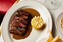 Sliced medium-rare steak with brown sauce on a white plate, served with a half lemon and a glass of wine nearby at Grand Brasserie.