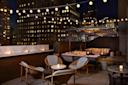 A stylish rooftop lounge at night with cozy seating, round tables, a marble-topped bar, string lights overhead, and city skyscrapers in the background. Candles on tables create a warm, inviting atmosphere.