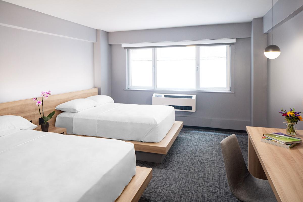 A modern hotel room with two double beds, a large window, a desk with a chair, a vase of flowers, and neutral-colored walls and carpet. The room is bright and simply decorated.