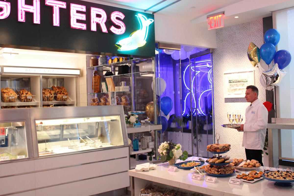 A man in a white coat stands with a tray of champagne flutes in a bakery. The display case is filled with pastries, and a table with assorted baked goods is in the foreground. Balloons and flowers decorate the room. Neon "FIGHTERS" sign above.