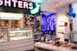 A man in a white coat stands with a tray of champagne flutes in a bakery. The display case is filled with pastries, and a table with assorted baked goods is in the foreground. Balloons and flowers decorate the room. Neon "FIGHTERS" sign above.