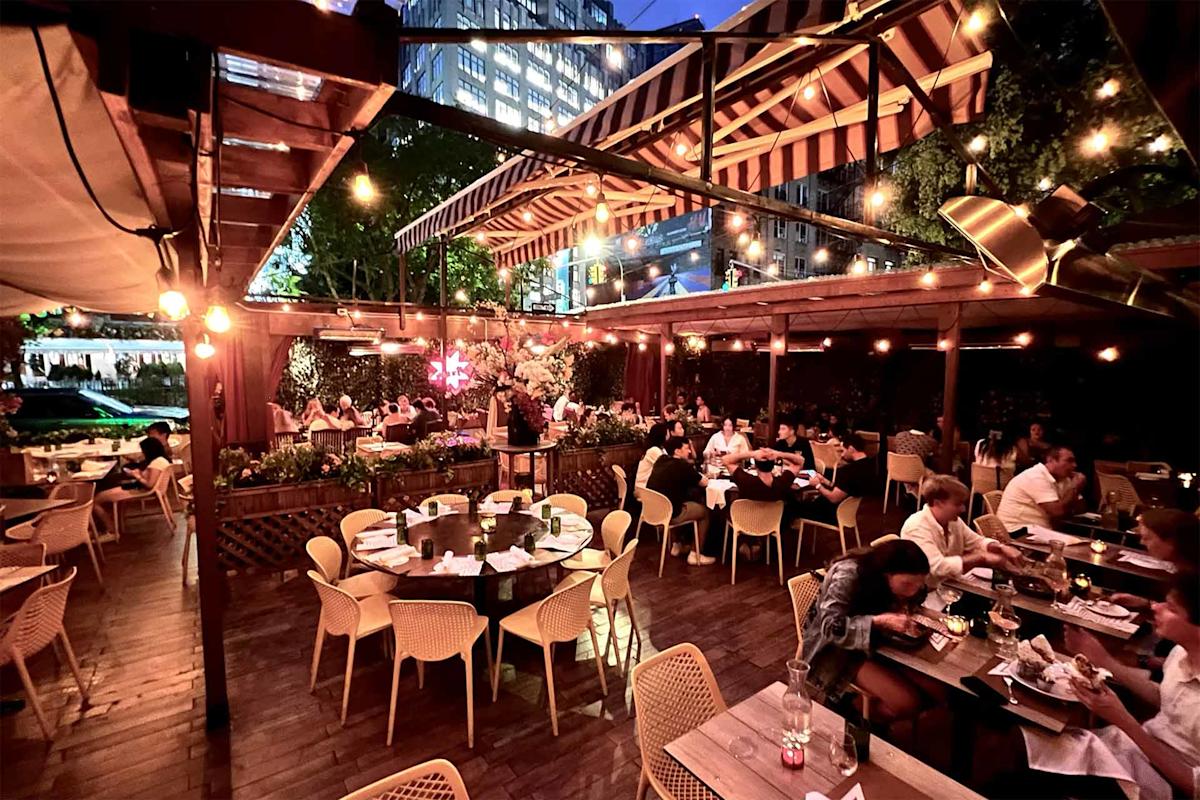 People dining at an outdoor restaurant patio in the evening, with string lights overhead and tall buildings visible in the background. The atmosphere is lively and cozy, with wooden decor and greenery around.