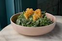 A bowl of kale salad topped with two crispy parmesan cheese crisps, served in a light pink bowl on a marble table with a teal wall in the background.