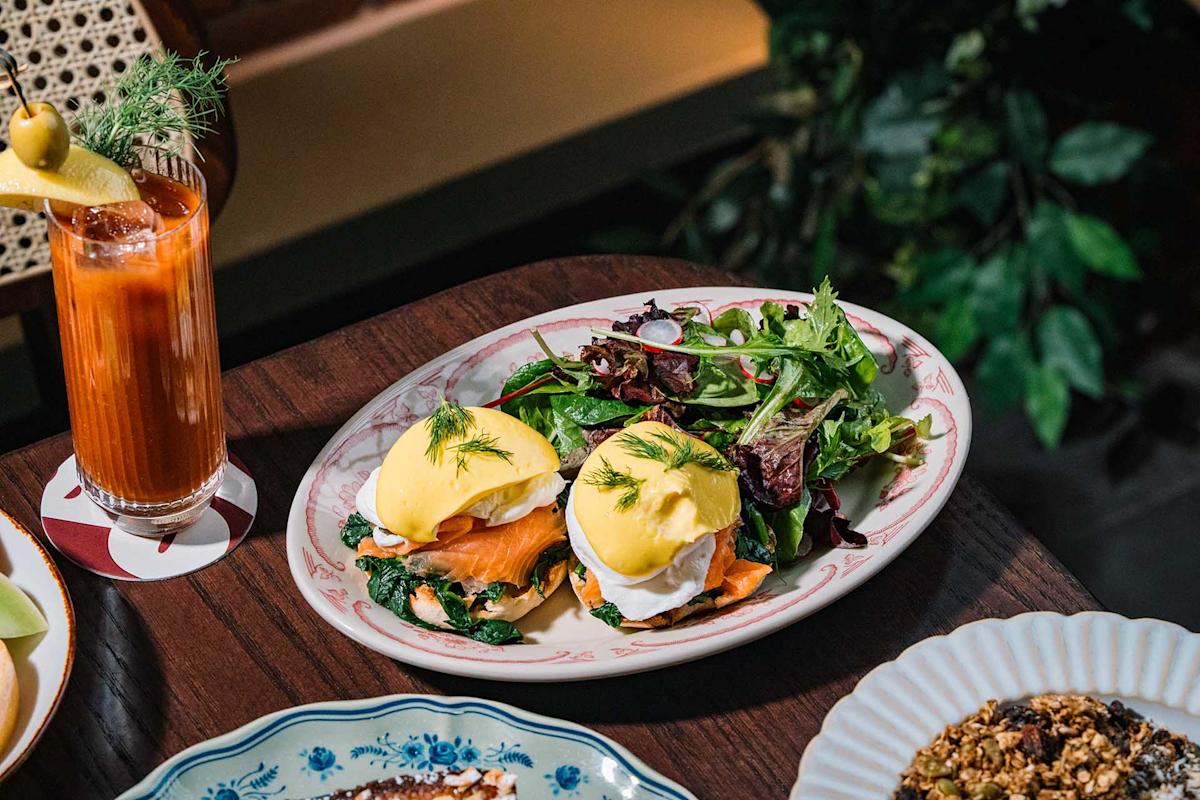 Plate with two eggs Benedict on smoked salmon and spinach, topped with hollandaise sauce and dill, served with a mixed green salad. A Bloody Mary cocktail sits nearby on the wooden table.
