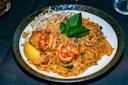 A plate of pad thai with shrimp, garnished with a basil leaf and crushed peanuts. Bean sprouts and a lemon wedge are on the side, adding a fresh touch to the dish. The noodles are golden brown and well-seasoned, served on a dark-rimmed plate.