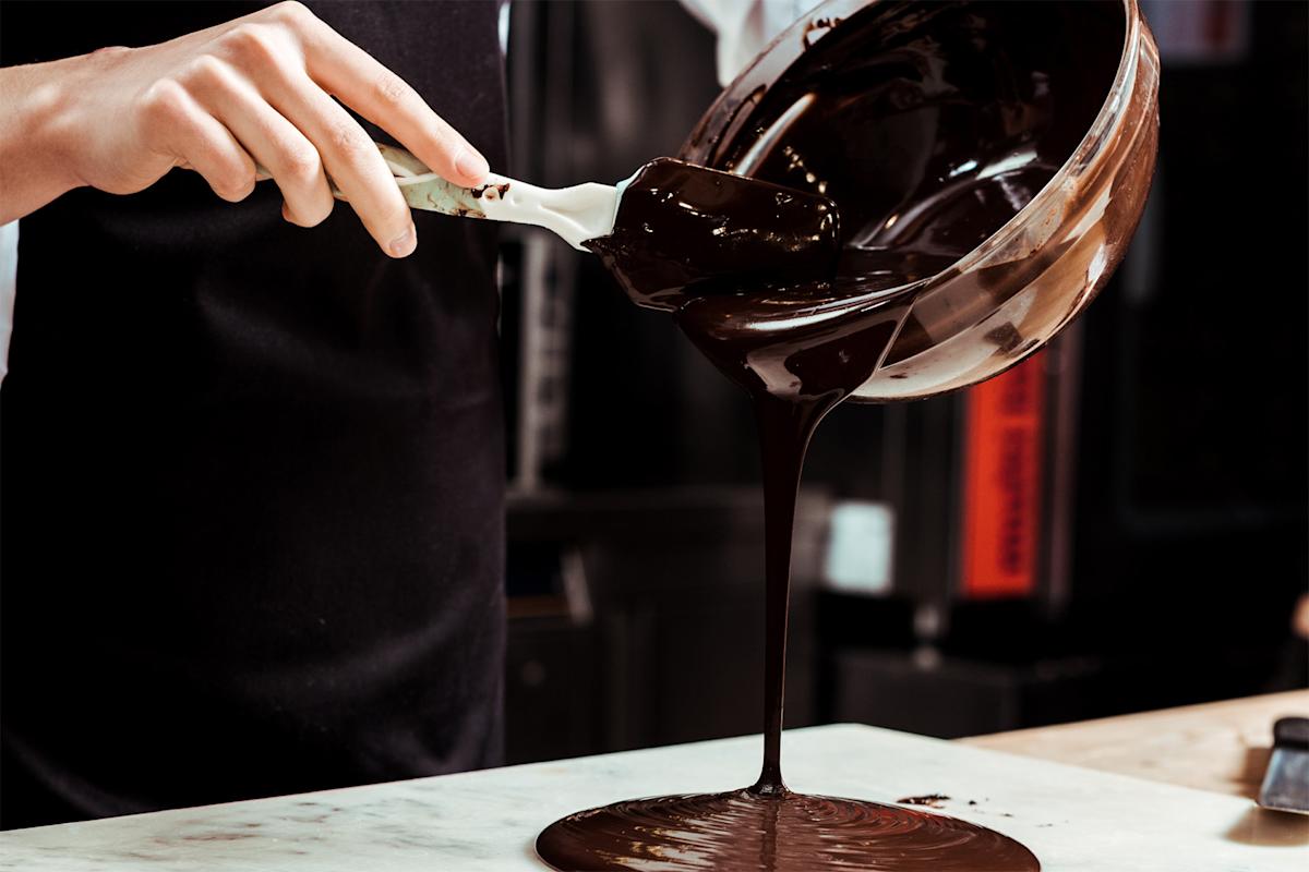 A person pours melted chocolate from a mixing bowl onto a marble surface, using a spatula. The scene is close-up, focusing on the flowing chocolate and the person’s hands.