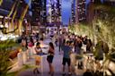 People gather and socialize at a lively rooftop bar in a city at dusk, with lights from tall buildings in the background and greenery lining the terrace. Some guests sit or stand at tables, enjoying drinks and conversation.