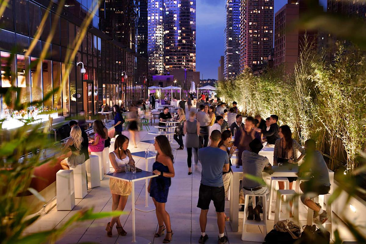 People gather and socialize at a lively rooftop bar in a city at dusk, with lights from tall buildings in the background and greenery lining the terrace. Some guests sit or stand at tables, enjoying drinks and conversation.