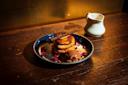 A blue ceramic plate with a stack of pancakes topped with chocolate ice cream and a chocolate stick. Rose petals are scattered around the pancakes. A small white pitcher sits in the background on a wooden table.
