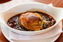 A golden-brown roasted duck leg served on a bed of lentils with a dark, rich sauce and garnished with cherries in a white oval dish atop a folded white napkin.