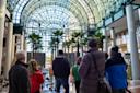 A group of people stand inside a large glass atrium with tall palm trees and holiday decorations, looking toward the bright, spacious entrance.