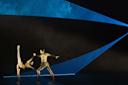 Two ballet dancers in gold costumes perform on stage; one holds the other’s hand as she stretches her leg high. Blue geometric lights create dramatic shapes in the dark background.