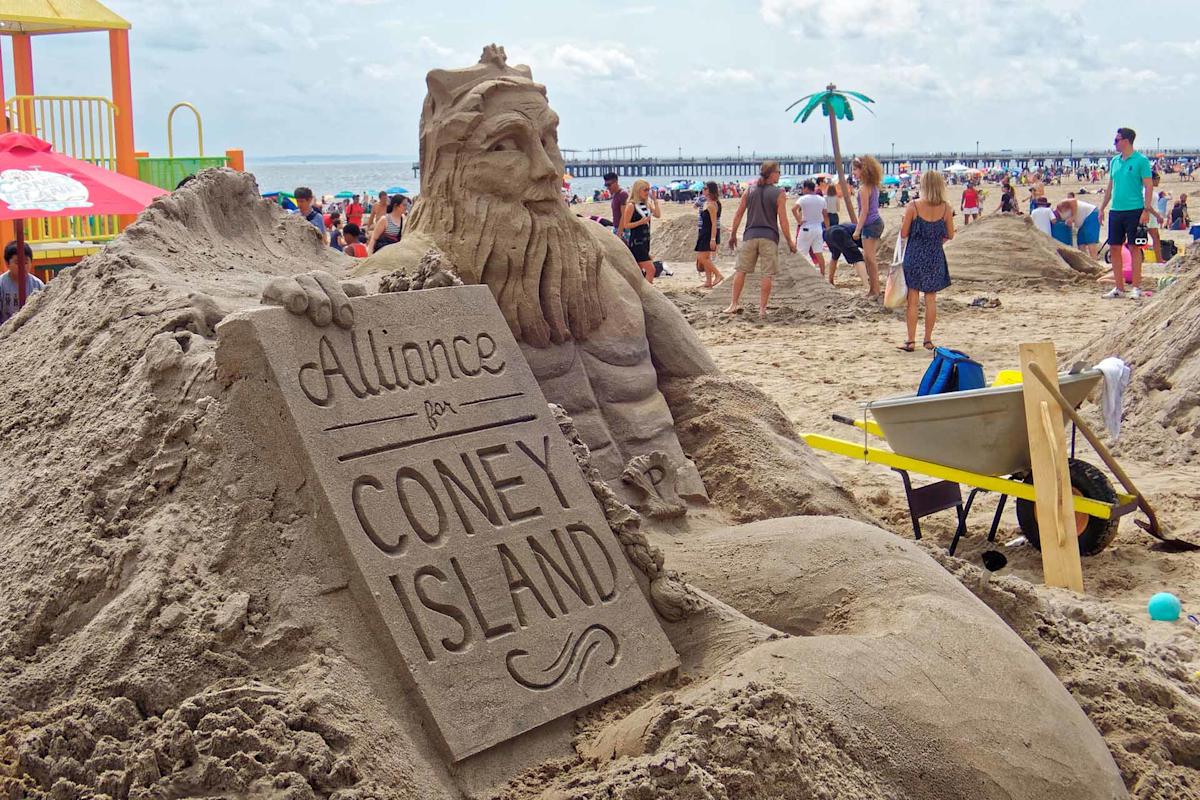 A detailed sand sculpture of a bearded figure holding a sign that reads “Alliance for Coney Island” stands on a crowded beach with people, a playground, and the ocean in the background.